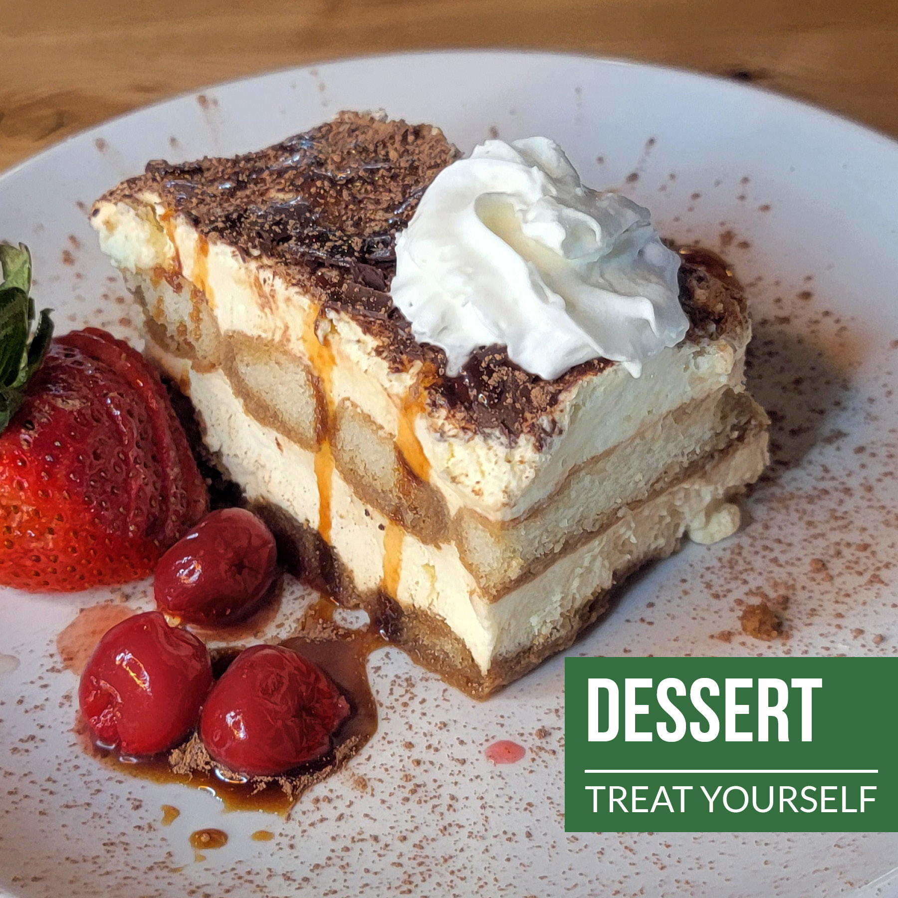 Tiramisu dusted with cocoa powder, topped with whipped cream, with a strawberry and cherries on the side